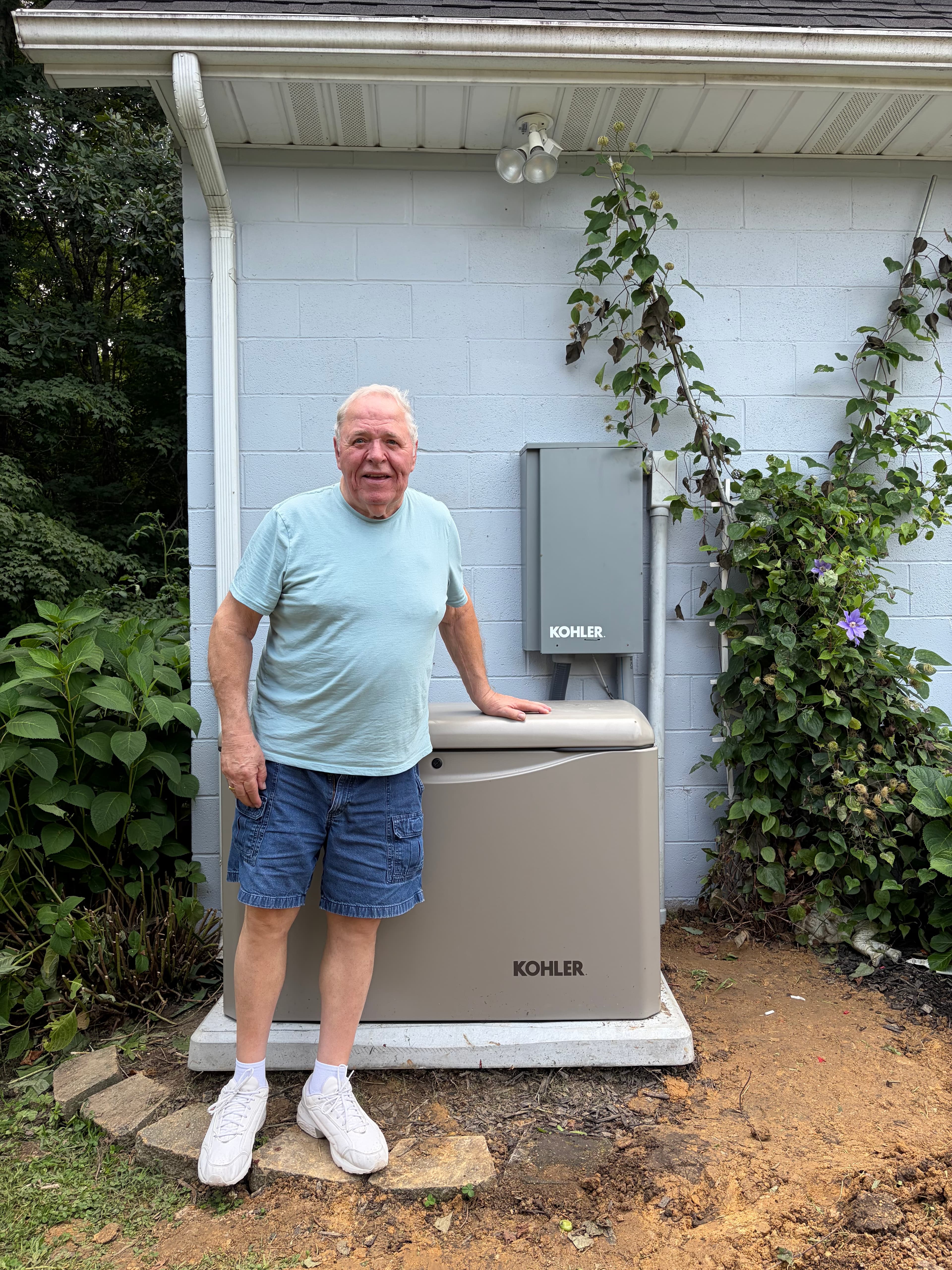 Homeowner posing beside a residential standby generator installed at the corner of a house